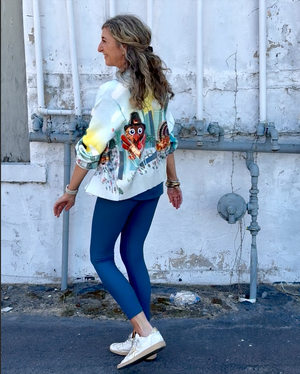 Woman wearing a colorful shirt with graphic designs and blue leggings standing against a white wall.
