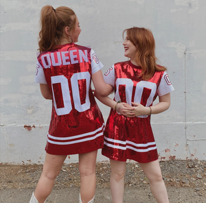 Two individuals linking arms wearing a red jersey dress front of a white wall.