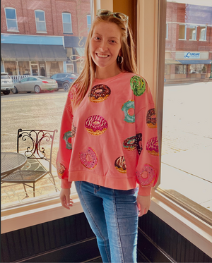 A individual wearing a pink sweatshirt with multi-colored donuts on it standing in front of a window.