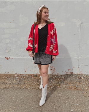 A person wearing a red cardigan with white and black text pattern, paired with a black top and white boots, standing against a white wall.