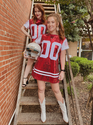 Two individuals wearing red and white full sequin jersey dresses with a football helmet accessory, standing on a staircase.