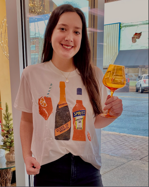 A person holding a glass of orange spritz wearing a white t-shirt with graphics related to spritz and a bottle of prosecco, and text that says 'Queen of Sparkles'.