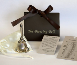 A silver bell sitting on a satin bag in front of a brown box labeled "The Blessing Bell" on a white backdrop. 