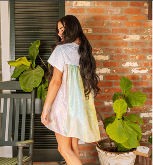A woman wearing a white iridescent sequin cape back dress with a rainbow sheen, standing in a sunny location with a brick wall and green foliage in the background.